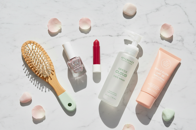 Flat lay of clean teen and tween beauty essentials on white marble — glitter nail polish, tinted lip balm, gentle face wash, SPF sunscreen, and bamboo hairbrush with pink and sage green accents — BodyWell1.com