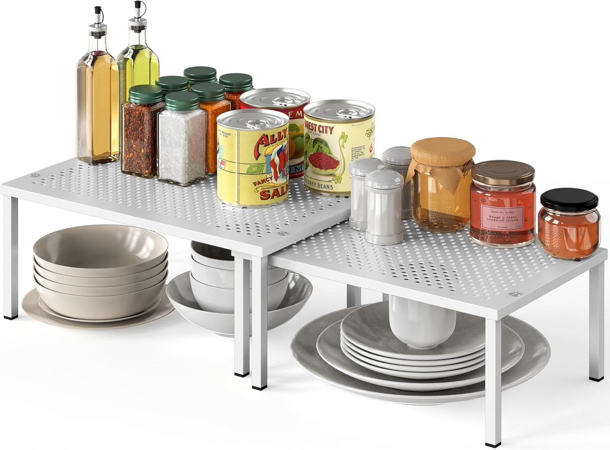 Two white perforated steel stacking shelves holding spices, cans, and dishes on a white background