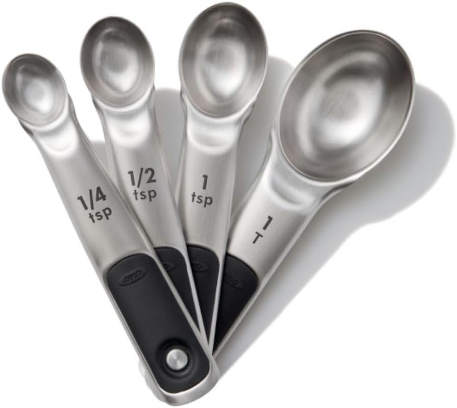 Four OXO stainless steel measuring spoons with etched teaspoon and tablespoon markings and soft black handles on a white background.